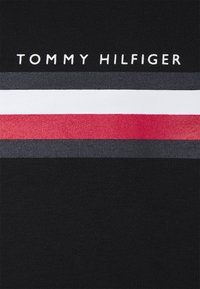 Logo Tommy Hilfiger umieszczone nad poziomymi paskami w ciemnoszarym, białym, czerwonym i ciemnoniebieskim kolorze na czarnym tle materiału.