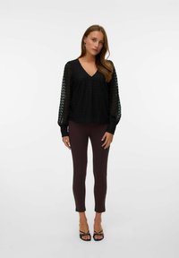 Zwarte, textuurrijke blouse met een V-hals en lange, doorschijnende mouwen, gecombineerd met aansluitende, donkerbruine leggings en zwarte strappy sandalen.