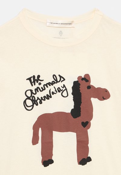 Κρεμ πουκάμισο με μία καφέ εικόνα αλόγου, μαύρη χαίτη και οπλές, μια μικρή καρδούλα στο στήθος και το κείμενο «The Animals Observatory» από πάνω.