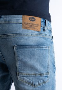 Pantalones vaqueros de mezclilla azul claro con acabado texturizado, que cuentan con una etiqueta de cuero marrón en la cintura trasera y dos bolsillos traseros.