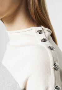 Ljusfärgad stickad tröja med hög krage, grå rand och dekorativa silvriga knappar längs axellinjen.