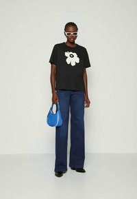 Black t-shirt featuring a large white flower graphic, paired with dark blue flared jeans and a small blue handbag. Model wears white sunglasses.