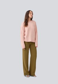 Pull oversize rose clair avec des manches longues associé à un pantalon large olive. Le mannequin se tient sur un fond gris uni.