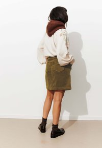 Jupe mini en velours côtelé kaki avec deux poches arrière, associée à un pull surdimensionné crème affichant des broderies florales et une capuche en tricot marron.