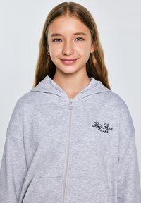 Siva zip-up kapuca s mehkim otipom, ki vsebuje vezeno logo "Big Star Jeans" v črni barvi. Ima kapuco in stranske žepe.