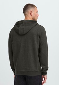 Man facing away wearing a dark gray hooded sweatshirt and black pants against a plain light background.