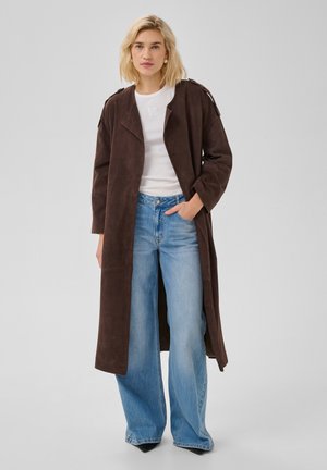 Blonde Frau steht, trägt einen langen dunkelbraunen Mantel, weißes T-Shirt, hellblaue weite Jeans und schwarze spitze Schuhe, Hand in der Tasche.