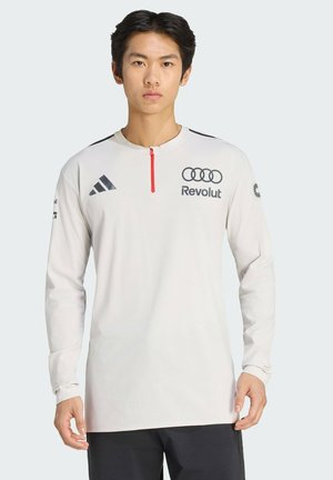 Junger Mann trägt ein langärmliges weißes Sportshirt mit den Logos von Adidas, Audi und Revolut, steht vor einem hellgrauen Hintergrund.