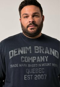 Námořnická modrá košile s dlouhým rukávem a vybledlým šedým textem "DENIM BRAND COMPANY" a detaily o místě a roce založení. Bavlněný materiál.