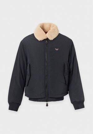 Homme portant une casquette noire, une veste bomber noire avec un col en peau lainée beige, un sweat-shirt noir avec logo, un pantalon noir et des chaussures marron foncé, debout.