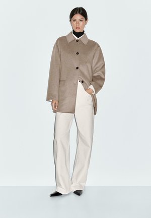 Manteau en mélange de laine beige avec un col structuré, des boutons noirs et des poches latérales, associé à un pantalon large crème et des chaussures noires à bout pointu.
