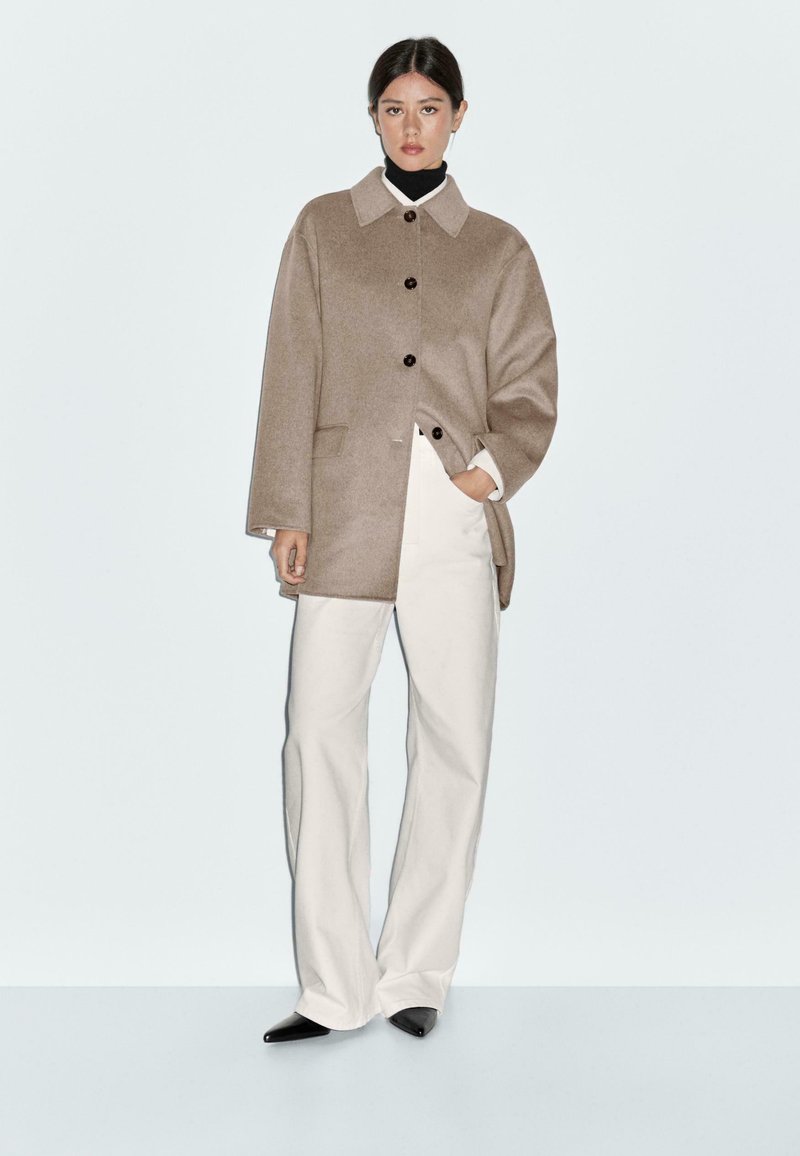 Beige wool-blend coat with a structured collar, black buttons, and side pockets, paired with cream wide-leg trousers and black pointed shoes.