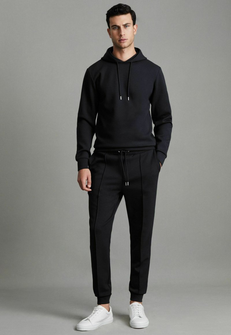Hanorac negru cu șireturi, asortat cu pantaloni jogger negri tapered, care au cusături laterale. Complețat cu pantofi sport albi pentru contrast.