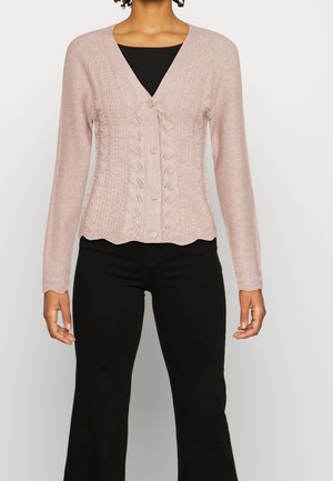 Kvinde iført en lyserød strikket cardigan med guldknapper over en sort top og sorte bukser, stående foran en ensfarvet baggrund.