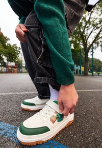 Baskets blanches avec des accents verts, une semelle texturée et un graphisme coloré. Portées avec un jean foncé et un pull en maille vert dans un cadre extérieur.