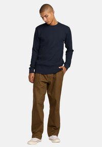 Marineblaues, geripptes Langarmshirt, kombiniert mit braunen, gerade geschnittenen Hosen und weißen Sneakers. Das Shirt hat einen runden Halsausschnitt und eng anliegende Bündchen.