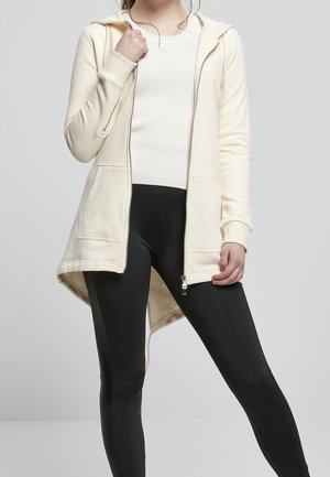 Femme portant un sweat à capuche crème zippé sur un top blanc côtelé et un legging noir, debout devant un fond gris clair uni.