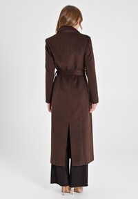 Cappotto lungo, marrone, con cintura e collo a punte, dotato di una fessura centrale sulla schiena e texture liscia. Indossato sopra pantaloni larghi scuri.