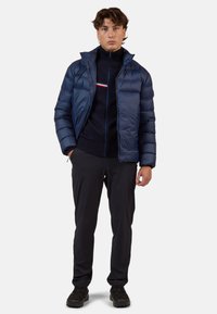 Giubbotto imbottito navy con chiusura a zip, dotato di un colletto alto e di un design imbottito. Indossato sopra un maglione scuro con dettagli a righe orizzontali.
