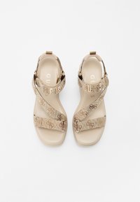 Beige Sandalen mit Kreuzriemen, die ein Blumenmuster und das "GUESS"-Logo zeigen. Glatter, flacher Sohlenrand mit einem verstellbaren Knöchelriemen.