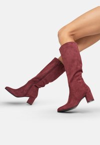 Botas de ante burdeos hasta la rodilla con un tacón cuadrado, puntera redondeada y detalle de cremallera trasera, que presentan una textura suave y un diseño ajustado.