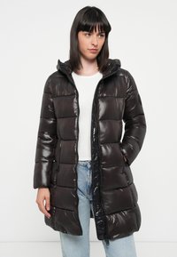 Sort pufferjakke med en skinnende finish, quiltet design, frontlynlås og hætte. Har sidelommer og en afslappet pasform. Bæres over en hvid top.