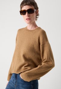Kašmirna pletena pulover s okroglo ovratnikom, sproščenim krojem, dolgimi rokavi in rebrastim robom, v kombinaciji z oversized očali iz želvovine.