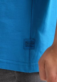 Camiseta de algodón azul con una etiqueta que dice "Petrol Ind" cerca del dobladillo, con mangas cortas y una textura suave.