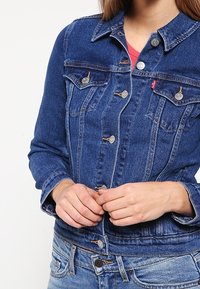 Džínová bunda v tmavě modré barvě s uzávěrem na knoflíky, dvěma náprsními kapsami a klasickým límcem. Dlouhé rukávy s detaily na manžetách.