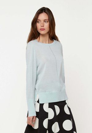 Femme aux longs cheveux bruns portant un pull bleu clair et une jupe noire à gros pois blancs, regardant sur le côté.