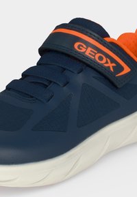 Gros plan d’une basket bleu marine avec doublure orange, lacets et une sangle Velcro marquée GEOX, dotée d'une semelle blanche.