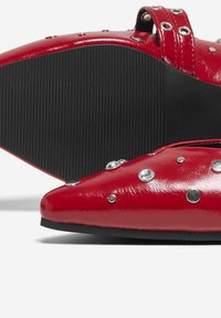 Zapatos de charol rojos con punteras afiladas, decorados con tachuelas metálicas y una suela de goma negra texturizada. Cuentan con una correa en el tobillo.