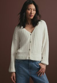 Vit rutig cardigan med V-hals, utrustad med fem dekorativa knappar och långa ärmar, ihop med blå blekta jeans. Slät textur.