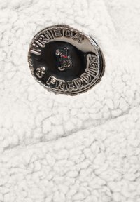 Wit fleece stof met een gestructureerd oppervlak heeft een ronde zilveren badge met de tekst "Frieda & Freddies" en toont een hond met een rode halsband.