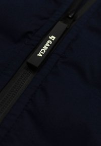 Närbild av svart dragkedjepull på mörkblått tyg med vit "GARCIA"-logotyp tryckt på dragkedjan.