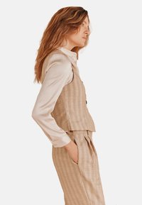 Beige textuurvest over een lichte crèmekleurige blouse, met een kraag en knoopdetails. Het model draagt ook bijpassende ruime broek.