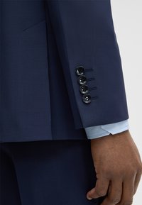 Dunkelblaues Anzugsmanschette mit vier schwarzen Knöpfen, hellblaues Hemdmanschette sichtbar und eine dunkelhäutige Hand entspannt an der Seite.