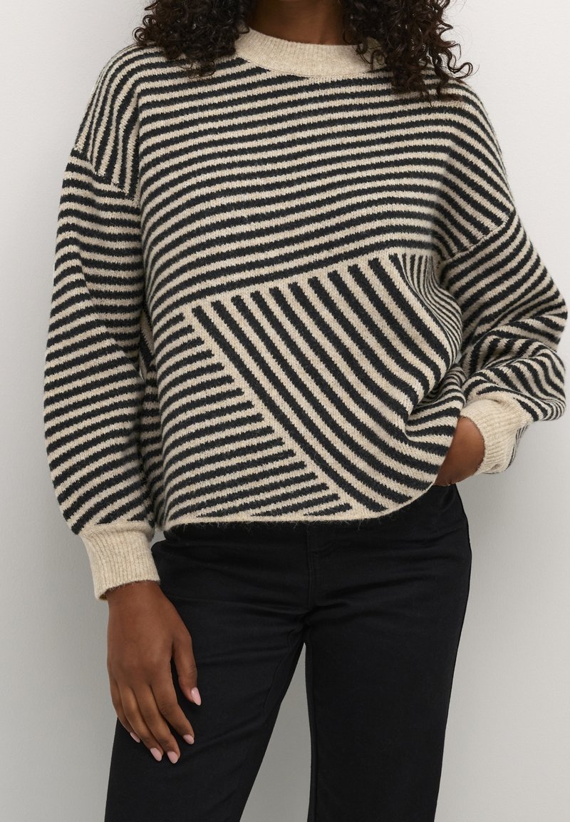 Femme portant un pull en laine beige et noir à rayures avec des motifs géométriques diagonaux et horizontaux, assorti à un pantalon noir.