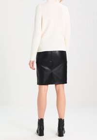 Cremfarbener Pullover mit hohem Kragen und strukturiertem Strick, kombiniert mit einem figurbetonten schwarzen Lederrock. Schwarze Stiefeletten runden das Outfit ab.