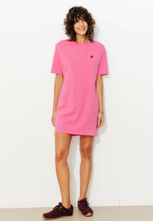 Femme aux cheveux bouclés portant une courte robe rose avec un petit cœur rouge sur la poitrine et des baskets foncées, debout contre un mur blanc.