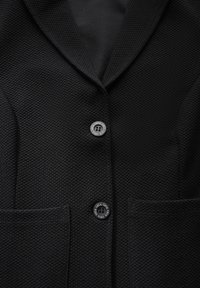 Černý texturovaný blazer s klopovým límcem, který má dvě náprsní kapsy a dvě zaoblené knoflíky s detaily značky.