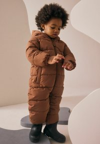 Brązowy pikowany kombinezon zimowy z kapturem, pikowaną teksturą i przednim zamkiem błyskawicznym. Do tego czarne gumowe buty; dziecko bawi się rękami.