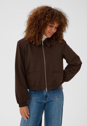 Femme aux cheveux bouclés portant une veste zippée marron foncé sur un jean bleu, regardant vers le bas et souriant avec une main sur la hanche.