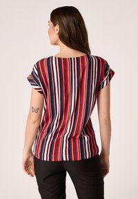 T-Shirt z krótkim rękawem z czerwonymi, białymi i granatowymi pionowymi paskami. Miękka tkanina o luźnym kroju i okrągłym dekolcie na plecach. Widoczny tatuaż na plecach.