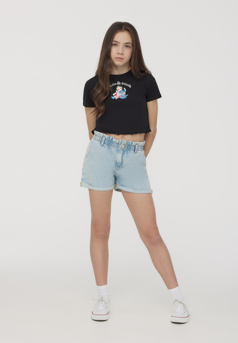 T-shirt nera corta con grafica "Lilo & Stitch", abbinata a pantaloni corti di jeans azzurri chiari con vita elastica e risvolti. Sneakers bianche.