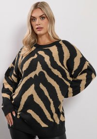 Blond kvinna som bär en överdimensionerad svart och beige zebra-randig tröja, poserar med en hand på höften mot en enkel vit vägg.