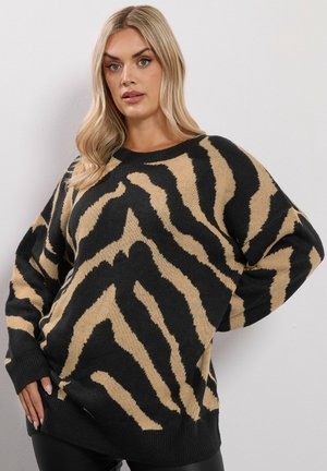 Blonde vrouw in een oversized zwart en tan gestreept zebra-trui, poseert met één hand op de heup tegen een plain witte muur.