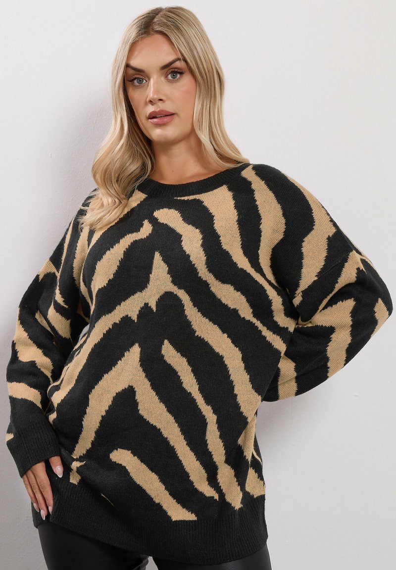 Blond kvinna som bär en överdimensionerad svart och beige zebra-randig tröja, poserar med en hand på höften mot en enkel vit vägg.