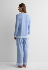 Frau steht barfuß und dem Gesicht zur Wand, trägt ein hellblaues langärmliges Oberteil und eine Hose mit weißer Spitzenverzierung an den Säumen und Bündchen.