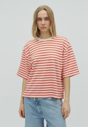 Jeune femme aux cheveux blonds mi-longs portant un t-shirt à manches courtes rouge et blanc rayé et un jean bleu clair sur un fond uni.